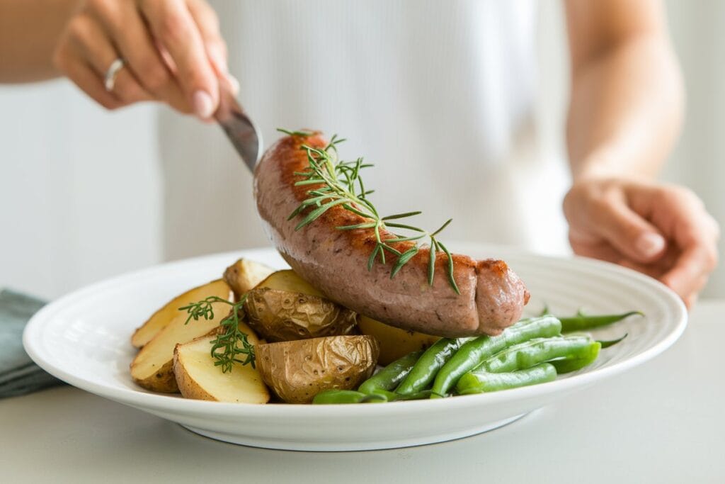 "A woman serving a perfectly cooked beef sausage dish, garnished with fresh herbs, atop a bed of sautéed onions and mushrooms. The plate is accompanied by a simple green salad with vinaigrette and a slice of crusty bread. The warm, inviting kitchen setting features a white tablecloth, enhancing the cozy atmosphere."