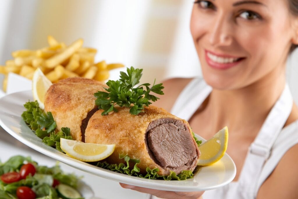 "A woman serving a perfectly cooked dish of Crispy Beef Milanesa, beautifully garnished with fresh herbs. The main dish is accompanied by classic sides, such as golden roasted potatoes and a fresh green salad. The scene is bright and inviting, with a warm ambiance that highlights the delicious meal."