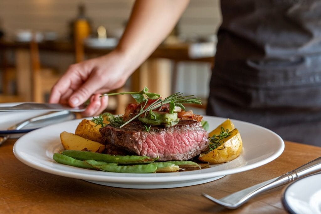 A woman serving a perfectly cooked Bavette steak, garnished with fresh herbs, on a white plate. The dish is accompanied by simple sides, including roasted potatoes and green beans. The background features a warm, inviting dining area with a wooden table and chairs.