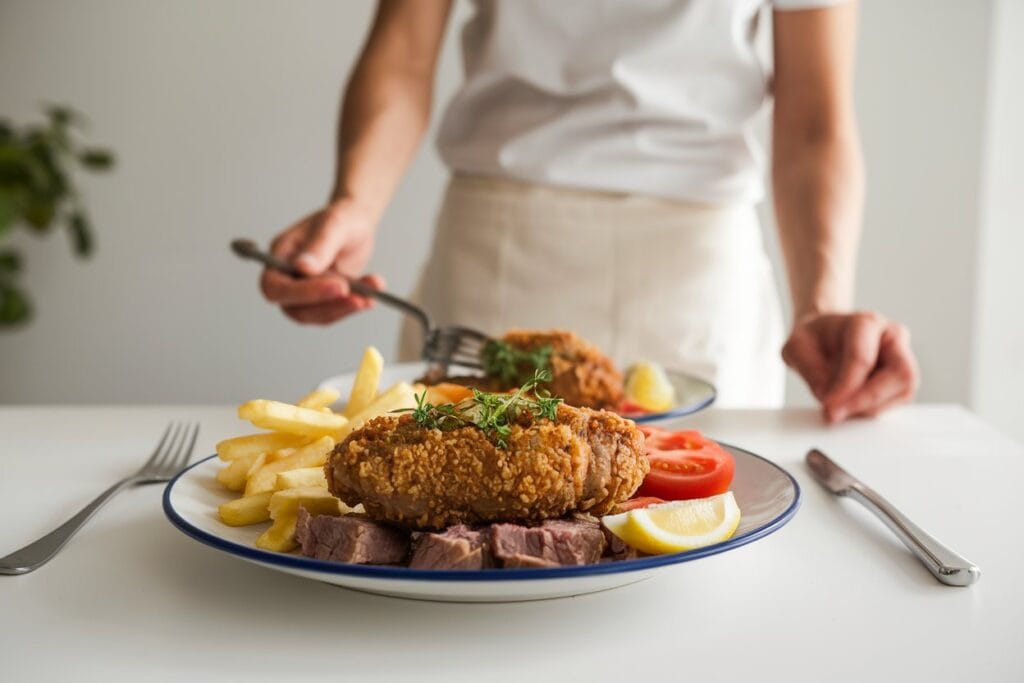 "A woman serving a perfectly cooked dish of Crispy Beef Milanesa, beautifully garnished with fresh herbs. The main dish is accompanied by classic sides, such as golden roasted potatoes and a fresh green salad. The scene is bright and inviting, with a warm ambiance that highlights the delicious meal."