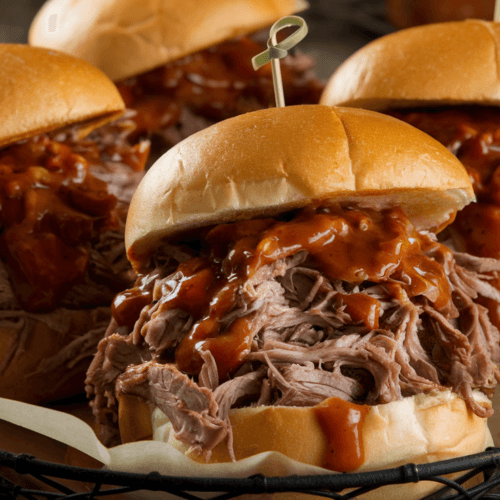 A close-up of pulled beef sandwiches served in a basket, overflowing with tender, juicy beef coated in a rich, tangy barbecue sauce. A toothpick holds each sandwich together, and the background features a rustic wooden surface for a warm, homey feel.