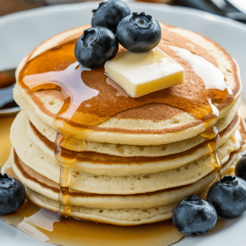 Fluffy gluten-free pancakes on a white plate, topped with fresh blueberries, a dollop of butter, and a drizzle of syrup. The golden-brown pancakes are neatly presented on a wooden surface with a few utensils in the background.