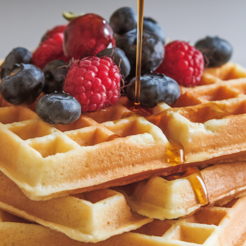 A stack of golden-brown gluten-free Belgian waffles with a light, fluffy texture and large holes. Topped with fresh berries and a drizzle of maple syrup, the waffles sit against a clean, simple background.